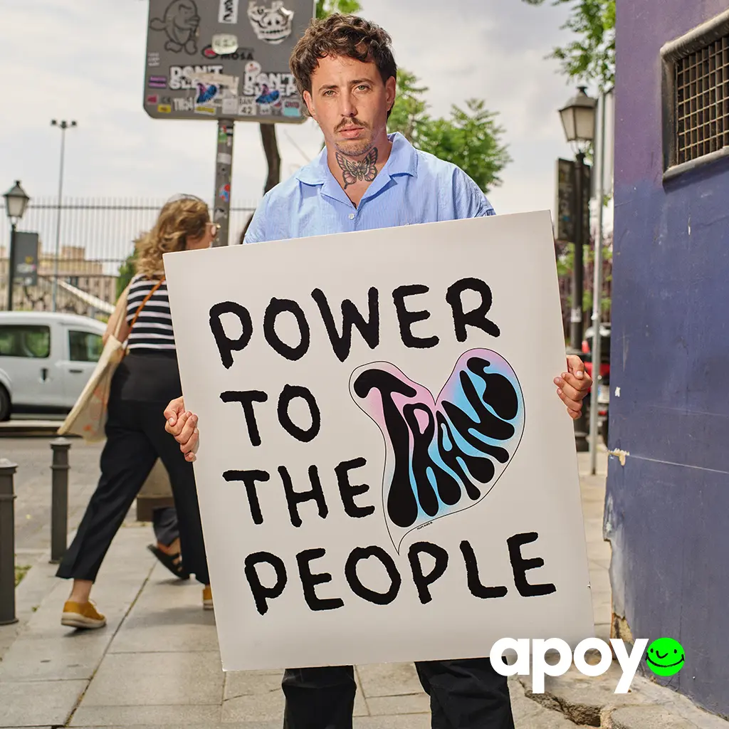 Hombre con camisa azul y tatuaje de mariposa en el cuello sostiene un cartel con el texto 'Power to the trans people' en una calle urbana. Imagen de campaña de la organización Apoyo.