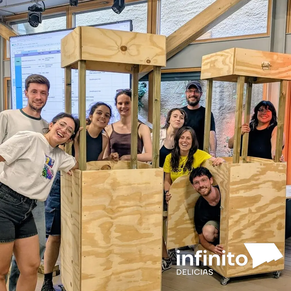 Nueve personas sonrientes posando entre estructuras de madera contrachapada en un espacio de trabajo creativo. Logo de Infinito Delicias en la esquina inferior derecha.